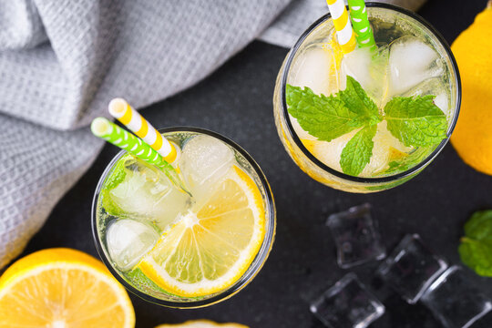Top view of refreshing homemade lemonade in glasses with fresh mint leaves, lemon slices, ice cubes and soda water and paper straws on a black background. Summer citrus drink, cold icy beverage.