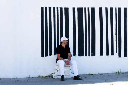 A tall, stylish, dark-haired, thin, 40-year-old white man with a beard is wearing white pants, black shoes, a black polo shirt., white cap.The background is a white wall with black barcode lines.
