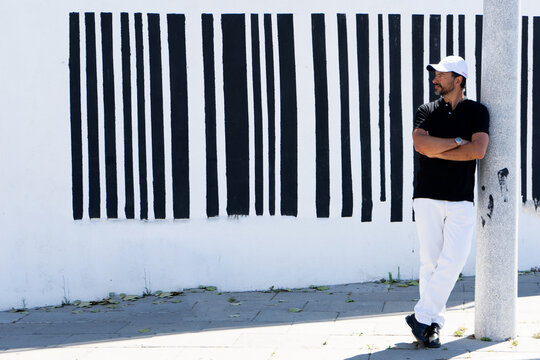 A tall, stylish, dark-haired, thin, 40-year-old white man with a beard is wearing white pants, black shoes, a black polo shirt., white cap.The background is a white wall with black barcode lines.