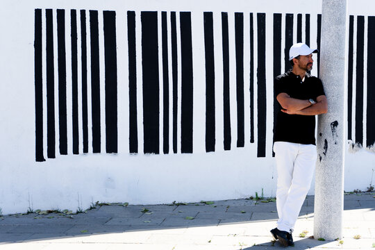 A tall, stylish, dark-haired, thin, 40-year-old white man with a beard is wearing white pants, black shoes, a black polo shirt., white cap.The background is a white wall with black barcode lines.