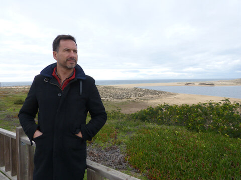 A dark-haired, bearded, white European man, wearing a winter coat, is relaxed in a beach area.