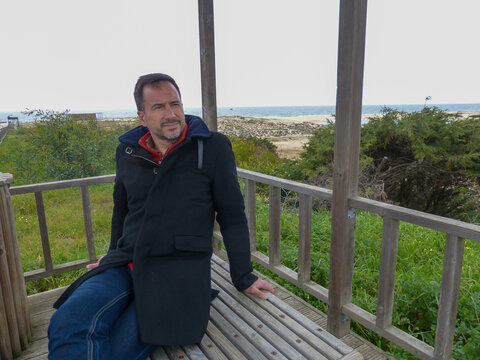 A dark-haired, bearded, white European man, wearing a winter coat, is relaxed in a beach area.