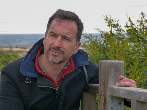 A dark-haired, bearded, white European man, wearing a winter coat, is relaxed in a beach area.