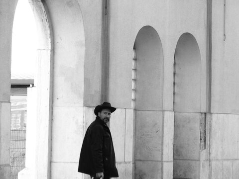 A stylish, dark-haired, thin, bearded white man of 40 years old, wearing a cowboy hat, moves with a trolley suitcase
