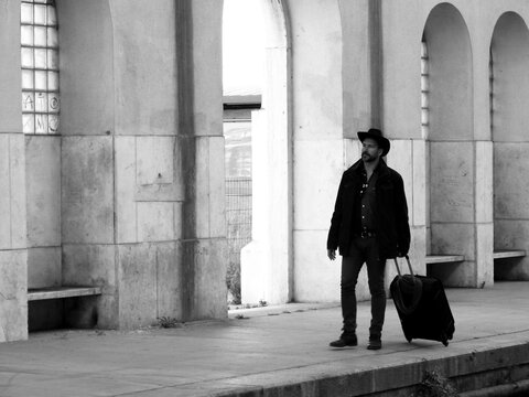 A stylish, dark-haired, thin, bearded white man of 40 years old, wearing a cowboy hat, moves with a trolley suitcase
