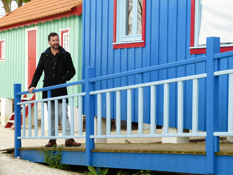 White man, dark-skinned, thin, 40 years old, casually dressed, posing against a backdrop of colorful wooden houses.