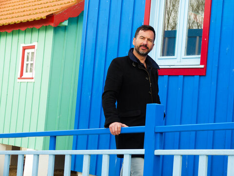 White man, dark-skinned, thin, 40 years old, casually dressed, posing against a backdrop of colorful wooden houses.