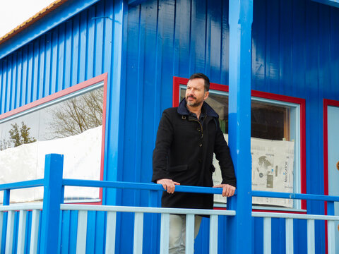 White man, dark-skinned, thin, 40 years old, casually dressed, posing against a backdrop of colorful wooden houses.