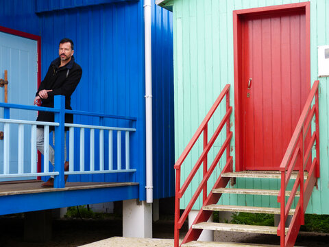 White man, dark-skinned, thin, 40 years old, casually dressed, posing against a backdrop of colorful wooden houses.