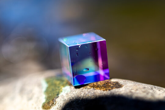 Color shifting glass cube rests on a river stone in bright sun. Iridescent purple and teal faces glow while tiny droplets cling to the surface and the stream behind dissolves into bokeh.
