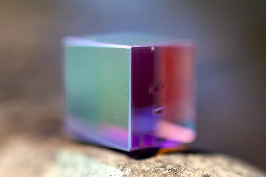 Iridescent glass cube rests on a rough stone with creamy bokeh and soft rainbow glow. Polished faces catch light and color creating a calm geometric mood in a quiet outdoor setting