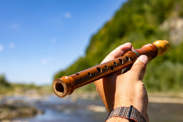 Perspective view of a wooden recorder held toward the river and sky. Sunlit details along the tube highlight tone holes craftsmanship and the timeless charm of acoustic wind instruments © Chornogirskyi