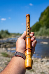 A hand firmly holds a polished wooden recorder beside a flowing river. Smooth grain and round tone holes stand out against the soft landscape suggesting tradition craft and acoustic melody © Chornogirskyi