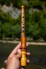 A hand holds a polished wooden flute with finger holes by a mountain river and green forest in bright daylight showcasing craftsmanship tradition and nature inspired music outdoors © Chornogirskyi