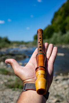 Wooden recorder rests across an open hand by a sunny river. Polished grain and finger holes show craftsmanship while the landscape blur adds a calm outdoor mood for folk music lovers