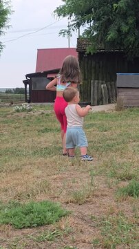 Two young siblings walking together on green grass outdoors in rural backyard near wooden barn during summer warm sunny day back view concept.