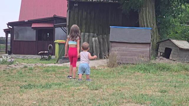 Two young siblings walking together on green grass outdoors in rural backyard near wooden barn during summer warm sunny day back view concept.