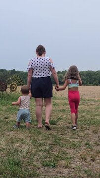 Mother walking hand in hand with her toddler boy and young daughter through a green rural field on a farm during summer day rear view.