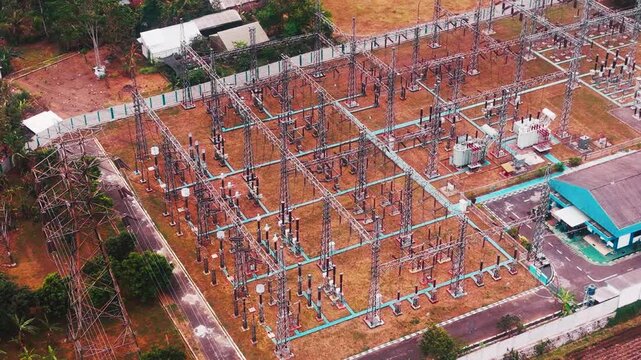 Aerial drone view of a large electrical substation featuring high voltage transformers, transmission towers, steel structures, switchgear, and power distribution equipment surrounded by dense urban bu
