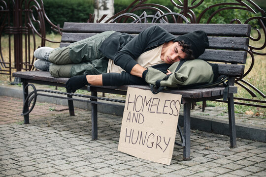 Homelessness and poverty crisis, young man sleeping on a park bench with a homeless and hungry sign