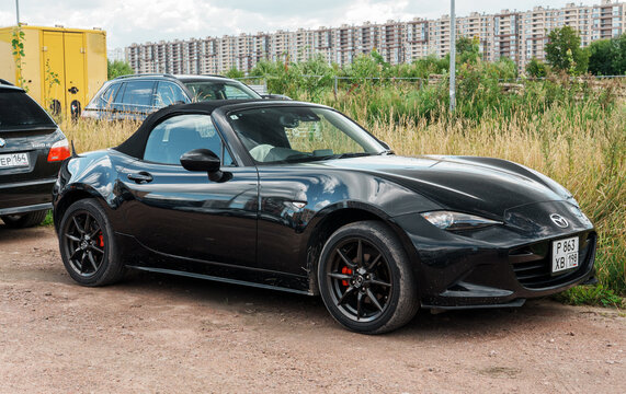 Exterior view of black Mazda MX5 Miata ND roadster convertible sports car parked on dirt road near green grass against residential city buildings and cloudy sky at summer day, auto photography. 