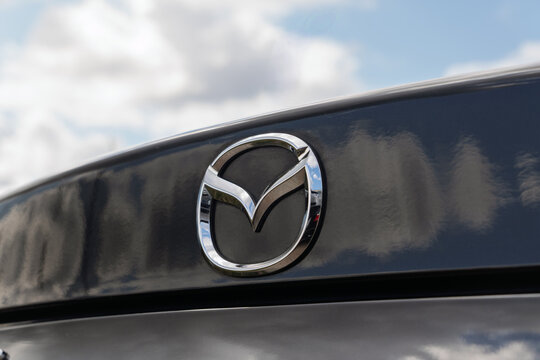 Close-up of silver chrome Mazda car emblem logo on glossy dark metallic trunk lid panel with reflection of cloudy sky at summer day, automotive brand sign identifier on vehicle rear exterior. 
