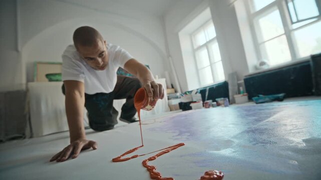 Rack focus shot of young adult man pouring red paint from jar onto large canvas on floor in slow motion. Footage suits art school promotions or creative industry campaigns