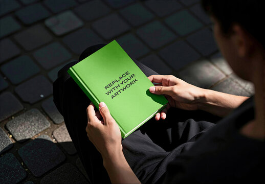 Book cover mockup held in hands on a cobblestone surface, realistic outdoor reading scene with natural sunlight, presentation template for print media and editorial design