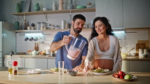 Man pouring smoothie pregnant wife kitchen. Couple enjoying healthy breakfast.