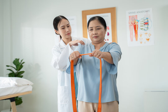 Caring young Asian women doctor smiling and encouraging elderly female patient doing arm stretching exercise with resistance band in clinic.Concept of physical therapy,rehabilitation,Physical Medicine