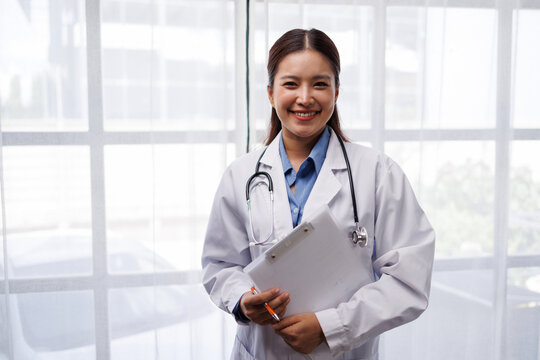 Asian female doctor standing in front of a bright window, wearing a white lab coat and stethoscope, smiling confidently while holding a clipboard and pen, representing professional healthcare