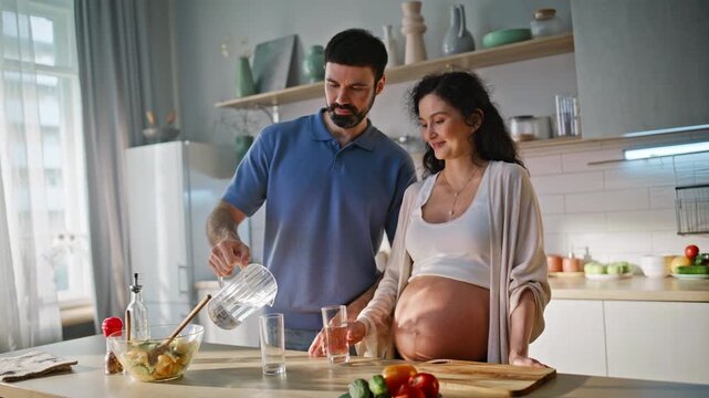 Man pouring water pregnant wife kitchen. Expectant parents clinking glasses.