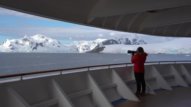 Female Photographer Antarctica Cruise Ship Passenger in Orange Coat on Deck Pulls up Telephoto Lens. Epic Adventure Travel Snow Covered Mountains