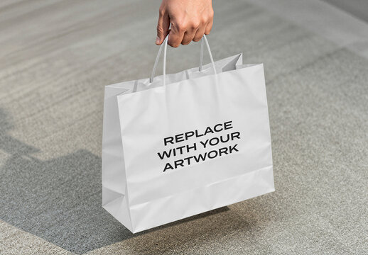 Paper shopping bag mockup held by hand on concrete surface, realistic packaging presentation for branding and advertising, outdoor urban environment with natural lighting