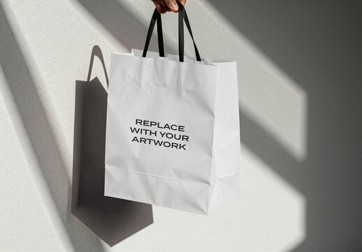 Paper shopping bag mockup with black handles held against a textured wall, realistic shadow and natural light, interior product presentation for branding, graphic design, print, advertising