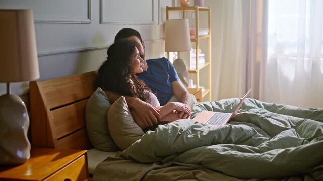 Pregnant couple relaxing bed browsing laptop. Expecting pair cuddling bedroom.