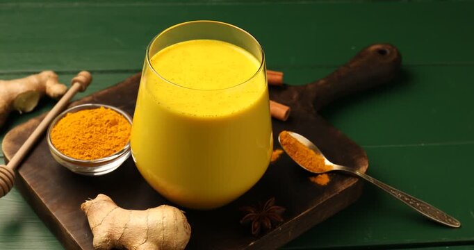 Golden milk turmeric latte in glass and ingredients on green wooden table, closeup. Camera moving right