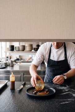 Professional pastry chef carefully plating an exquisite gourmet dessert with a spun sugar sphere in a modern restaurant kitchen