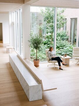 Woman sitting in a modern Scandinavian lounge with floor to ceiling windows overlooking a lush green garden courtyard