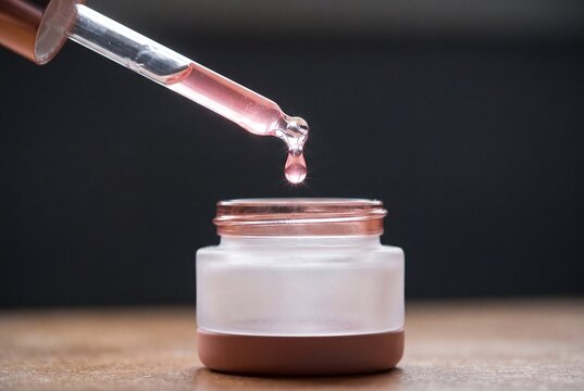 Pink cosmetic serum dripping from glass pipette into a skincare jar on wooden table with dark background