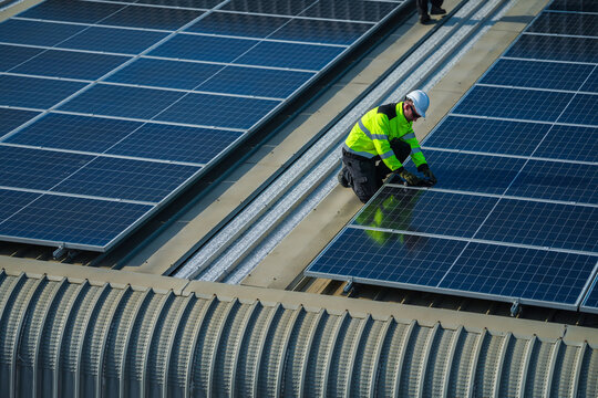 A photovoltaic technician wearing a safety vest and hard hat calmly crouches over the panels for a final evening test high on an industrial warehouse roof in a Thai city.