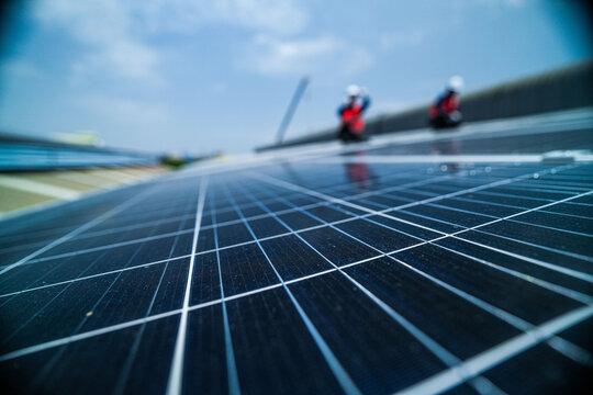 Two renewable energy specialists in hard hats and hi vis workwear quietly crouch pointing across the wet mirror like modules high on an industrial warehouse roof in a Thai city.