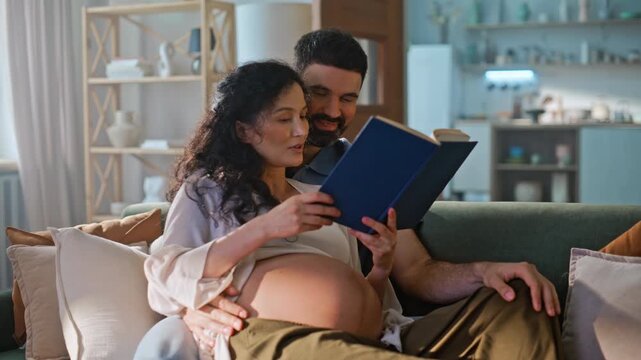 Expectant parents reading book sofa. Husband touching pregnant belly home.