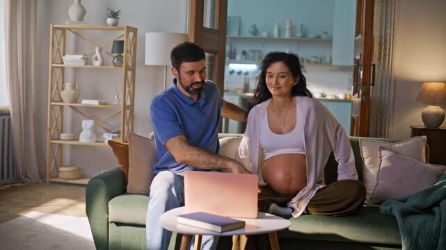 Expecting couple watching laptop home sofa. Pregnant woman husband together.