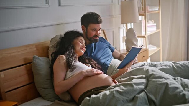 Expecting parents reading book bed. Man embracing pregnant wife cozy bedroom.