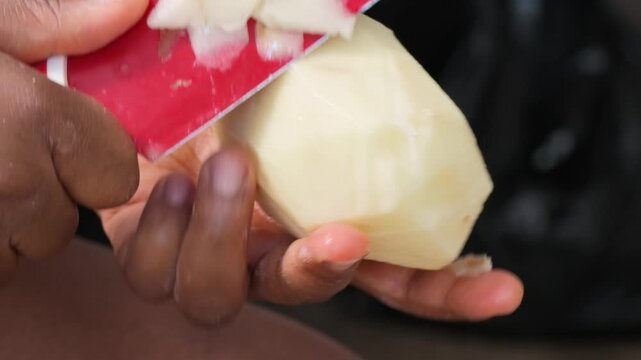 Hands scraping peeled raw potato with red kitchen knife close up