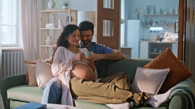 Couple expecting baby home sofa. Man touching pregnant belly holding tea cup.