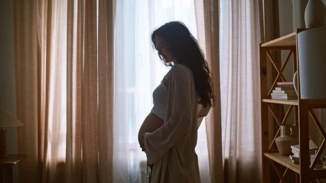 Expecting mother touching belly home. Backlit pregnant lady near window.