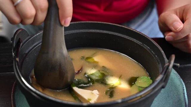 Person stirring hot miso soup with tofu and seaweed in ceramic bowl.