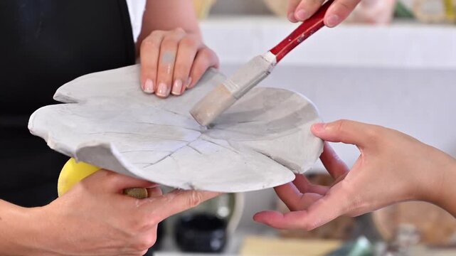 Pottery instructor applying grey liquid clay slip on ceramic leaf shape plate in studio.